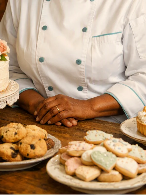 Carol Lynn Daley~Murray at her work table, hands clasped beside a rose-topped celebration cake, plates of chocolate chunk and sugar cookies, and a lemon meringue tart